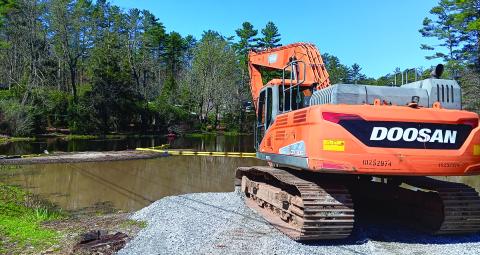 An excavator sits at the ready next to Mirror Lake where the yellow boundary lines for dredging have been put in place.
